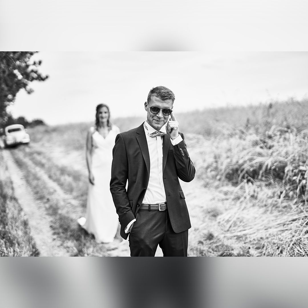 Marié en costume avec des lunettes de soleil sur un chemin, sa femme floue en arrière-plan